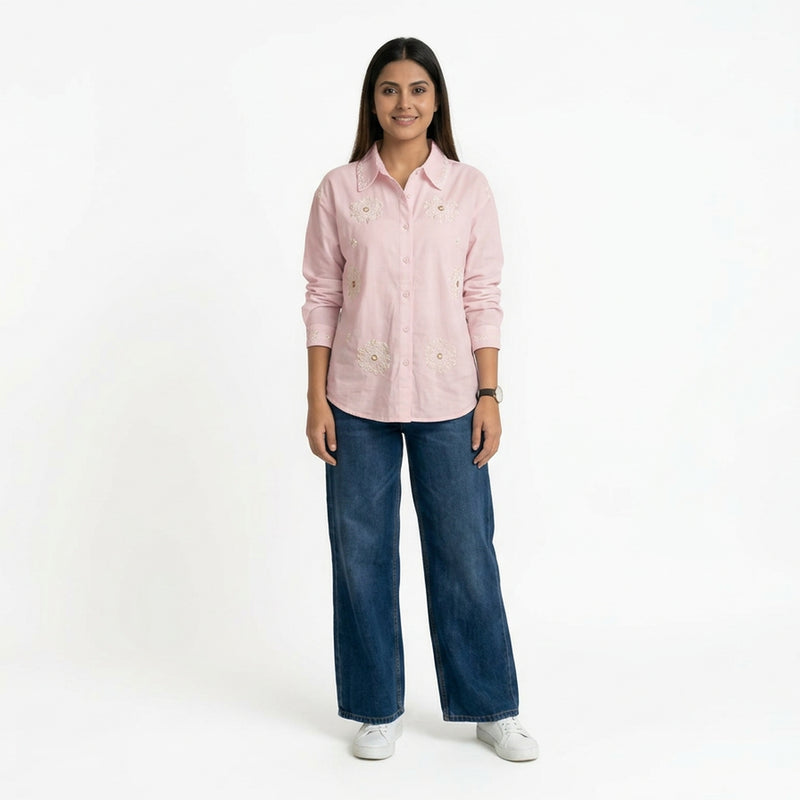 A woman wearing a light pink button-down shirt with floral embroidery, paired with blue wide-leg jeans and white sneakers.