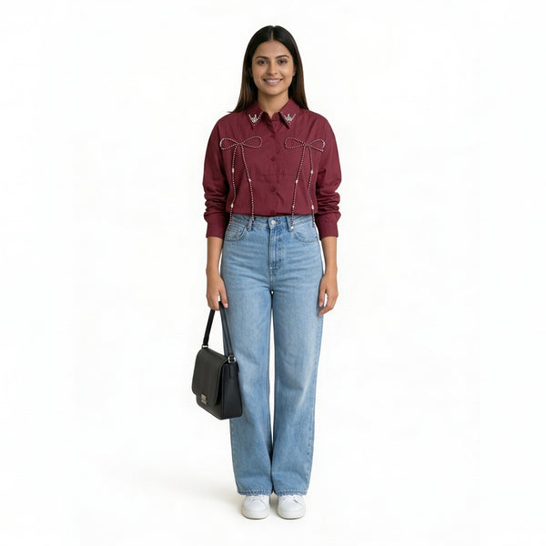 A woman wearing a maroon button-down shirt with bow embellishment details on the front, paired with light blue straight-le