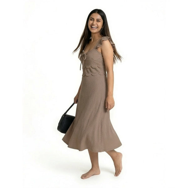 A smiling woman wearing a sleeveless brown midi dress with ruffled straps and a textured finish, walking barefoot while holding a structured black handbag.
