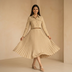Woman wearing a beige cropped jacket with a matching pleated midi skirt, posing in a minimal indoor setting.

