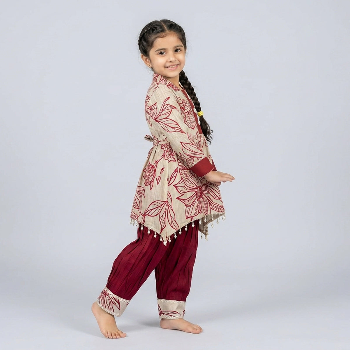 Young girl wearing a traditional outfit with a floral top and maroon pants on a light gray background