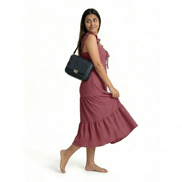 A woman wearing a sleeveless maroon midi dress with ruffled straps and a tiered, textured skirt, walking barefoot while carrying a black shoulder bag.