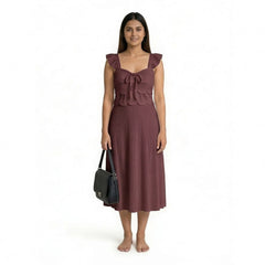 A woman wearing a sleeveless maroon midi dress with ruffled straps, scalloped detailing, and a textured finish, standing barefoot and holding a structured black handbag.