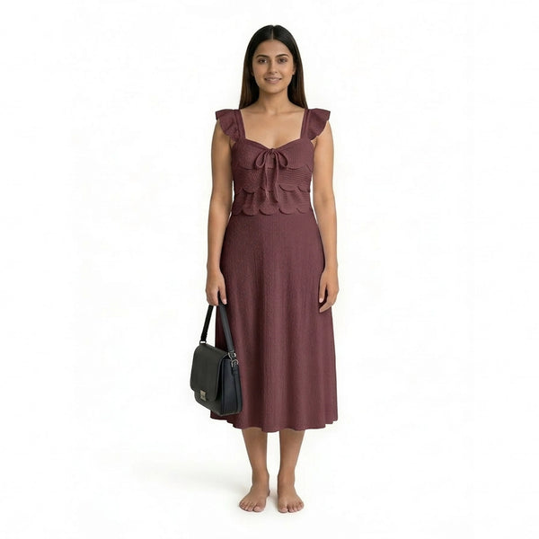 A woman wearing a sleeveless maroon midi dress with ruffled straps, scalloped detailing, and a textured finish, standing barefoot and holding a structured black handbag.
