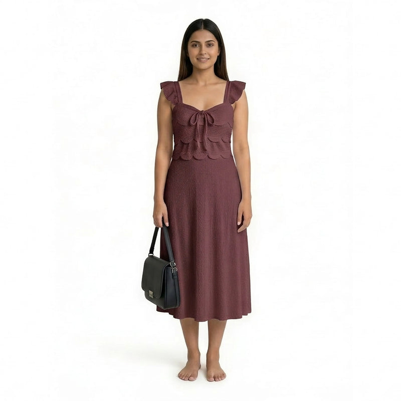 A woman wearing a sleeveless maroon midi dress with ruffled straps, scalloped detailing, and a textured finish, standing barefoot and holding a structured black handbag.