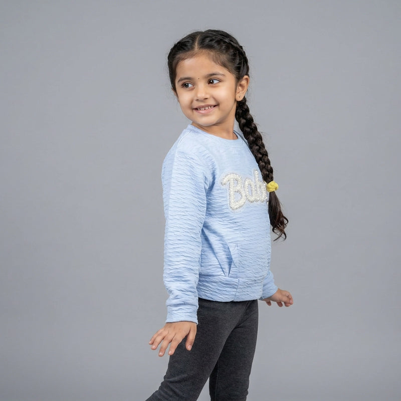Young girl wearing a light blue sweater with 'Bella' on it against a gray background