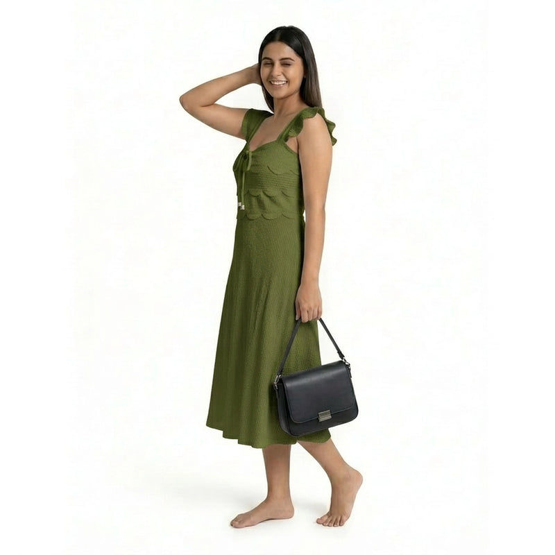 A smiling woman wearing a sleeveless olive green midi dress with ruffled straps and a textured finish, posing barefoot while holding a structured black handbag.
