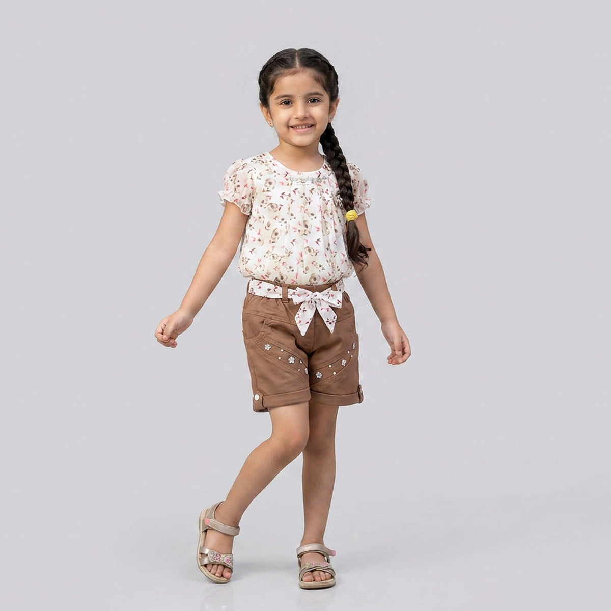 A young girl with a long braid is modeling a two-piece outfit consisting of a short-sleeve cream top with a small floral print and brown shorts with white embroidered details, set against a solid light gray background.