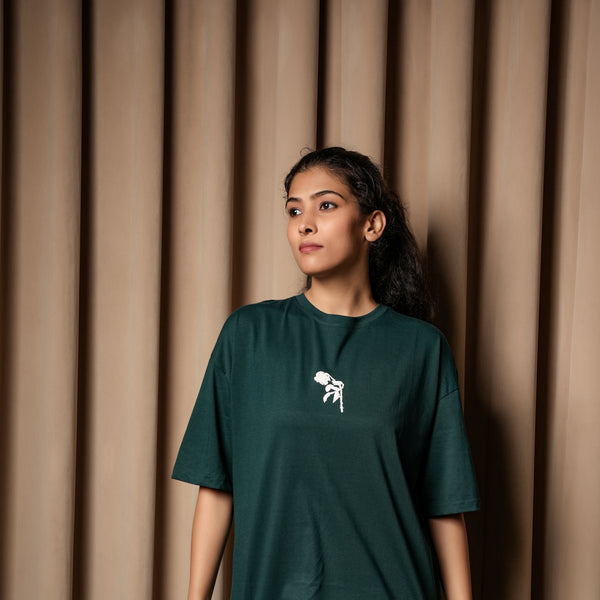 A woman wearing a dark green oversized T-shirt with a small white graphic on the chest, standing against a curtain backdrop.