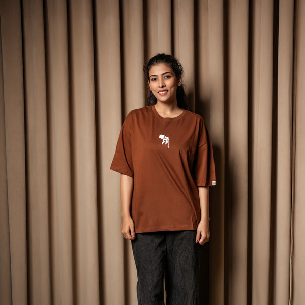 A woman wearing a brown oversized T-shirt with a small white graphic on the chest, paired with dark jeans, standing against a curtain backd

