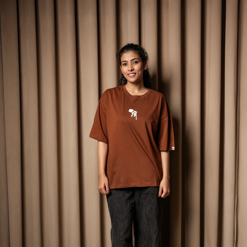 A woman wearing a brown oversized T-shirt with a small white graphic on the chest, paired with dark jeans, standing against a curtain backd


