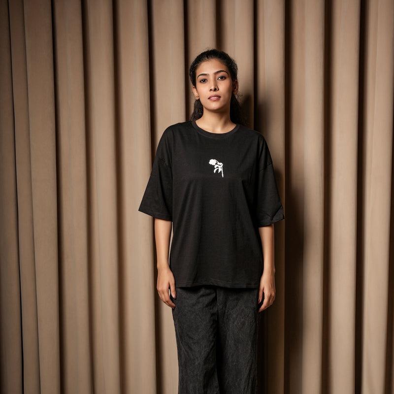 Woman wearing a black oversized T-shirt with a small white chest graphic, paired with dark jeans, standing against a beige curtain background.

