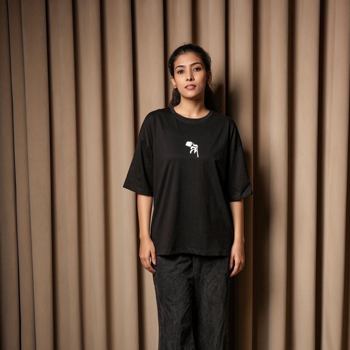 Woman wearing a black oversized T-shirt with a small white chest graphic, paired with dark jeans, standing against a beige curtain background.

