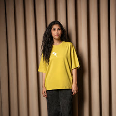 Woman wearing a mustard yellow oversized T-shirt with a small white chest graphic, paired with dark jeans, standing against a beige curtain background.