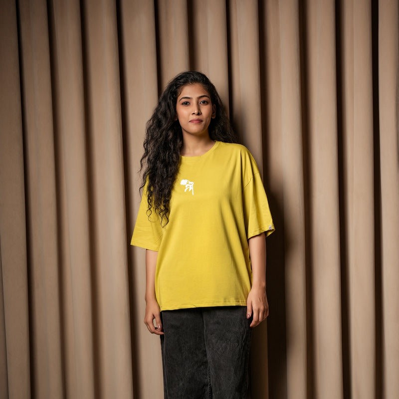 Woman wearing a mustard yellow oversized T-shirt with a small white chest graphic, paired with dark jeans, standing against a beige curtain background.