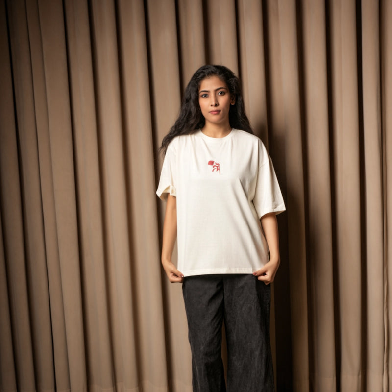 Woman wearing an off-white oversized T-shirt with a small red chest graphic, paired with dark jeans, standing against a beige curtain background.