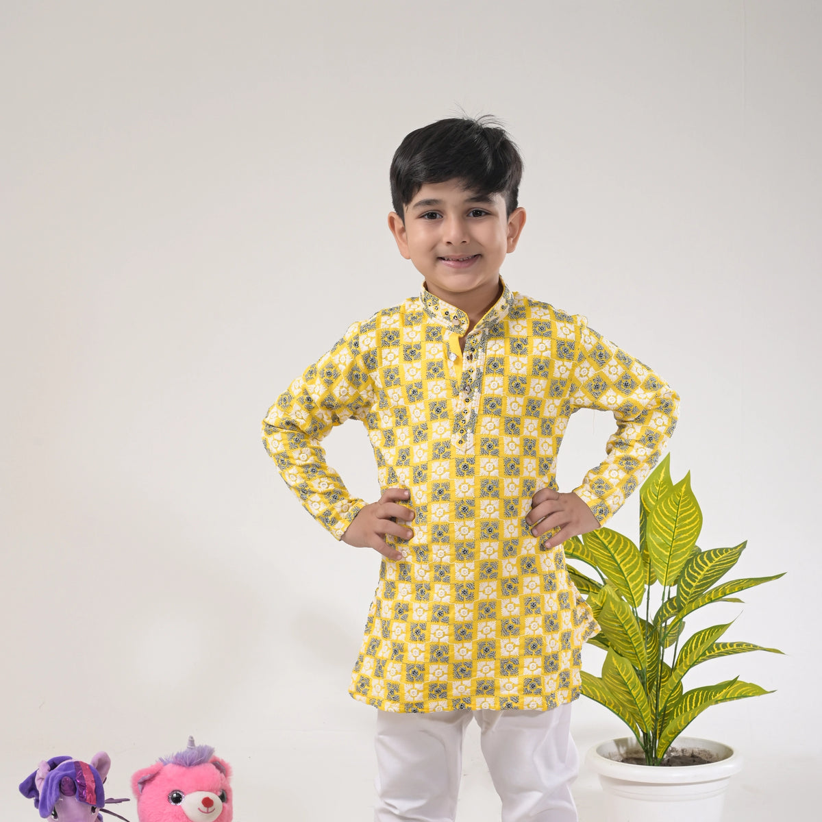 Child wearing a yellow patterned kurta with white pants, standing next to a plant and stuffed toys on a light gray background.