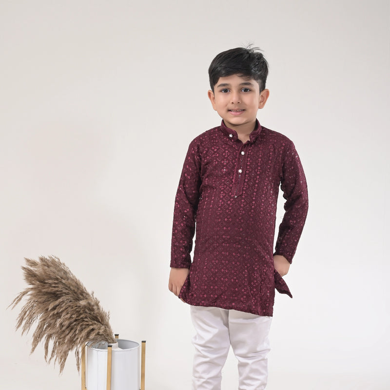 Child wearing a maroon kurta with white pants standing against a plain background