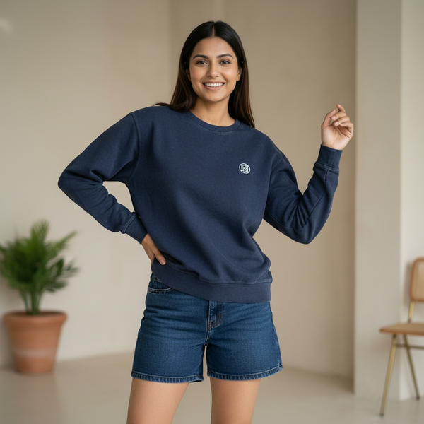 Woman wearing a navy blue sweatshirt with a minimal logo detail, paired with denim shorts, smiling and posing in a clean indoor setting with soft decor.

