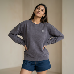 Woman wearing a grey sweatshirt with a small logo detail, paired with denim shorts, posing confidently in a minimal indoor setting.

