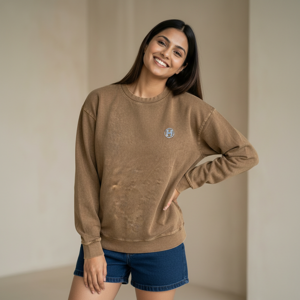 Woman wearing a brown sweatshirt with a subtle logo detail, paired with denim shorts, smiling and posing in a minimal indoor setting.

