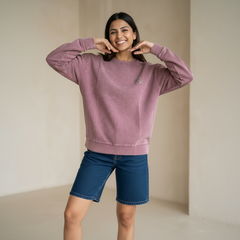 Woman wearing a dusty pink sweatshirt with a small logo detail, paired with denim shorts, smiling and posing playfully in a minimal indoor setting.

