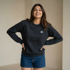 Woman wearing a black sweatshirt with a small logo detail, paired with denim shorts, smiling and posing in a minimal indoor setting.

