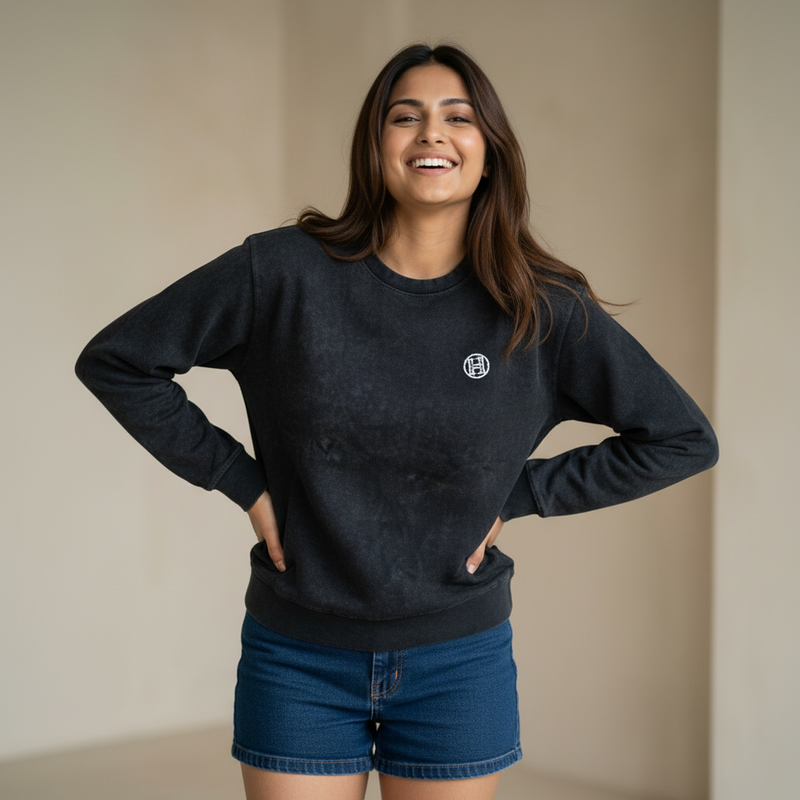 Woman wearing a black sweatshirt with a small logo detail, paired with denim shorts, smiling and posing in a minimal indoor setting.

