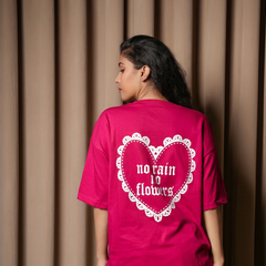 Back view of a woman wearing a bright pink oversized T-shirt featuring a white heart graphic with “no rain no flowers” text, standing against a beige curtain background.

