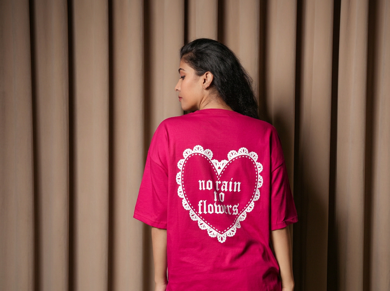 Back view of a woman wearing a bright pink oversized T-shirt featuring a white heart graphic with “no rain no flowers” text, standing against a beige curtain background.

